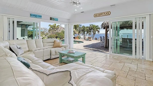 A spacious living room with a white sofa, a glass coffee table, and a ceiling fan. A sign reading "OUR HAPPY PLACE" is visible. - The Boat House · Riverfront · Heated Pool & Spa · Tiki Bar · Sleeps 10 (Cape Coral)