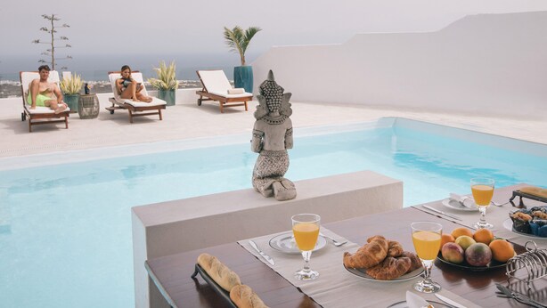 Een eetgedeelte bij het zwembad met een tafel gedekt voor het ontbijt, inclusief croissants en fruit, en twee ligstoelen met uitzicht op de oceaan.