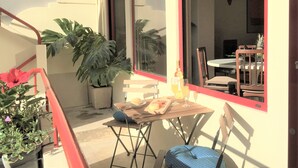 Un petit espace extérieur aménagé pour prendre les repas, avec une table et des chaises, des plantes en pot et une vue sur l’intérieur à travers une fenêtre.