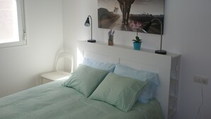 Une chambre à coucher comprenant un lit, une chaise, une table de chevet, une lampe et une étagère fixée au mur, sur laquelle sont posées deux plantes en pot.