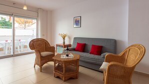 A living room with wicker furniture, a grey sofa with red cushions, and a glass table with a bottle and glasses.