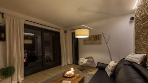 Een moderne woonkamer met een donkere bank, een houten salontafel met kaarsen, een vloerlamp en een schuifpui naar een balkon.