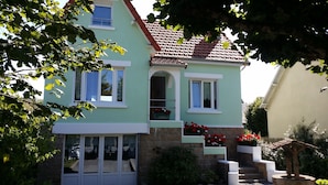 Une maison à deux étages, avec un toit recouvert de tuiles rouges, un balcon orné de fleurs rouges et un escalier en pierre menant à l’entrée.