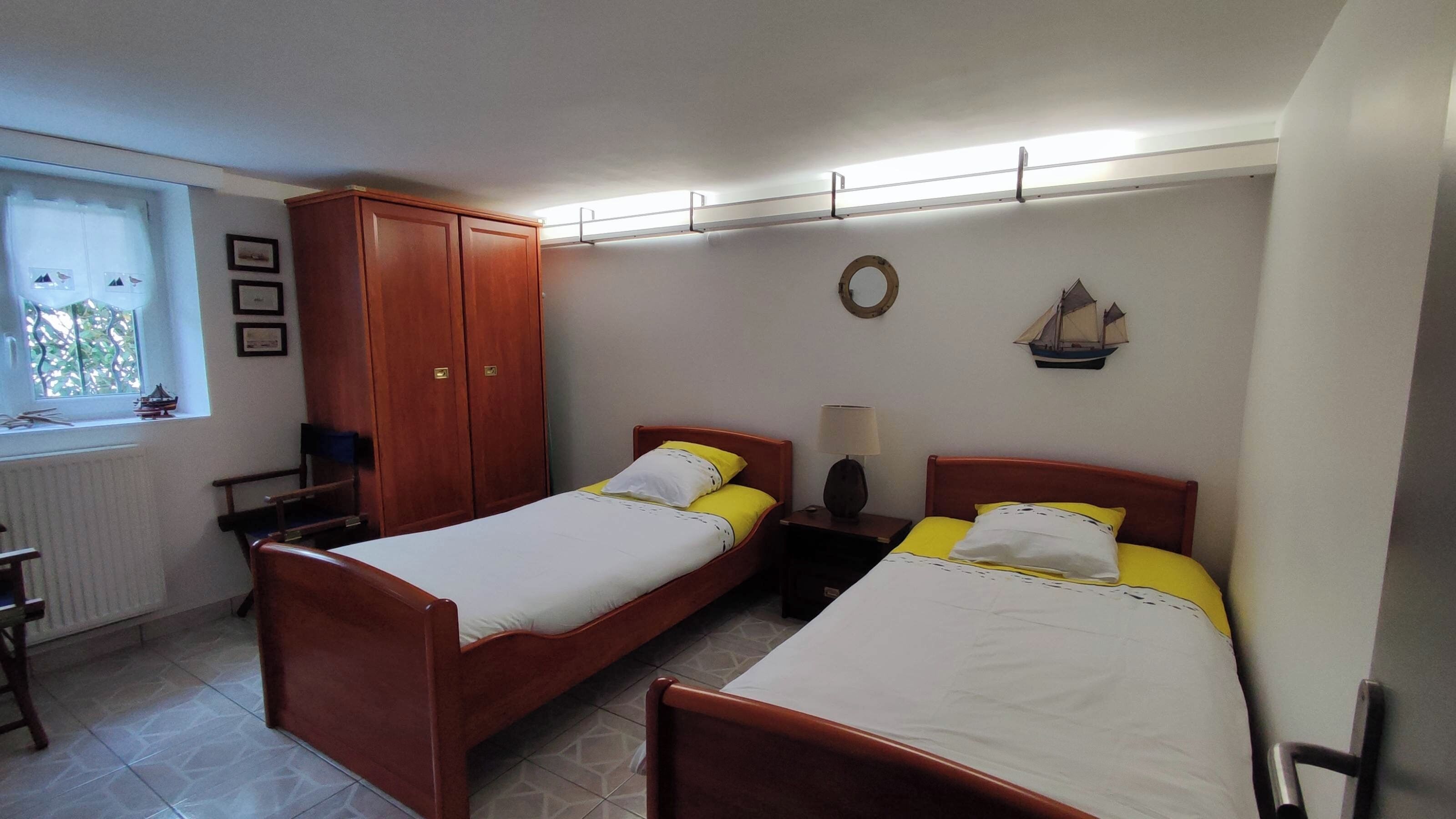 Une chambre avec deux lits en bois, une armoire, une petite table avec une lampe, une fenêtre donnant sur un paysage agréable et une horloge murale.
