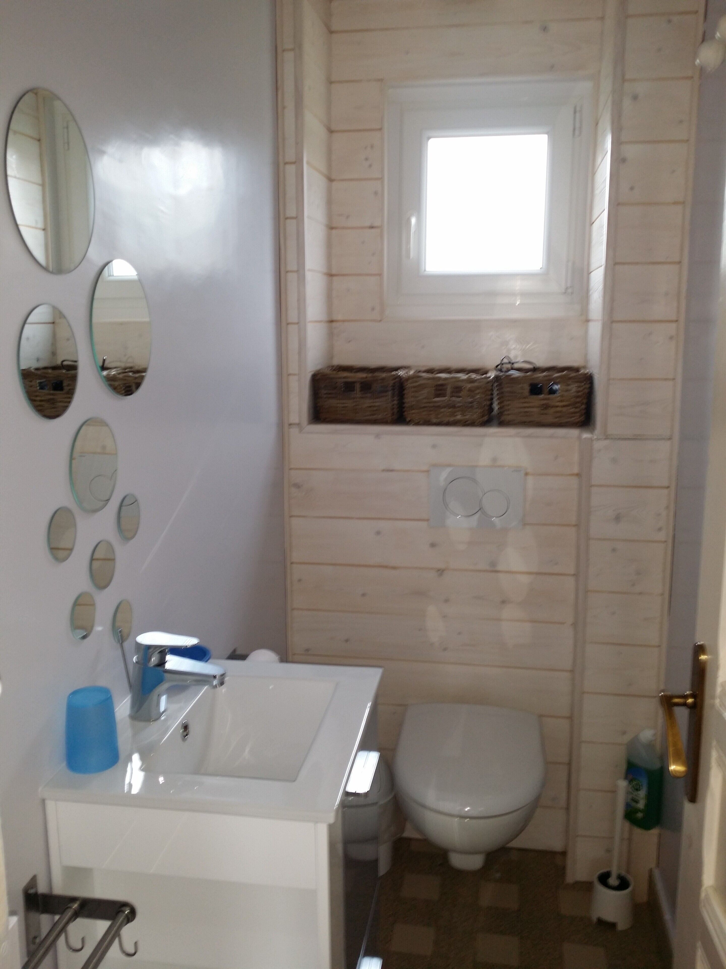 A bathroom with a white sink, a toilet, and a window above the toilet. There are several mirrors on the wall and baskets on the shelf above the toilet.