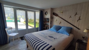 Une chambre avec un lit, une chaise, une fenêtre avec des rideaux, une étagère murale ornée de tableaux et un mur en bois décoré de motifs d’oiseaux.