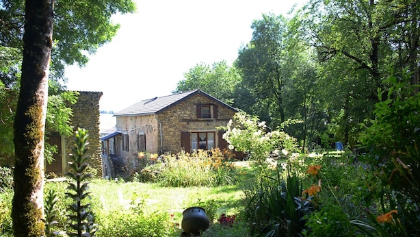 A stone house with a sloped roof, surrounded by lush greenery and flowering plants.