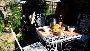 A metal table with a striped tablecloth, surrounded by white metal chairs. There are plates with bread, a bottle, and a thermos on the table.