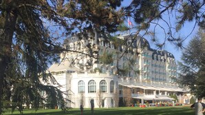 Un grand bâtiment historique à plusieurs étages, doté de fenêtres en arc de cercle, entouré d’une pelouse bien entretenue et d’arbres.