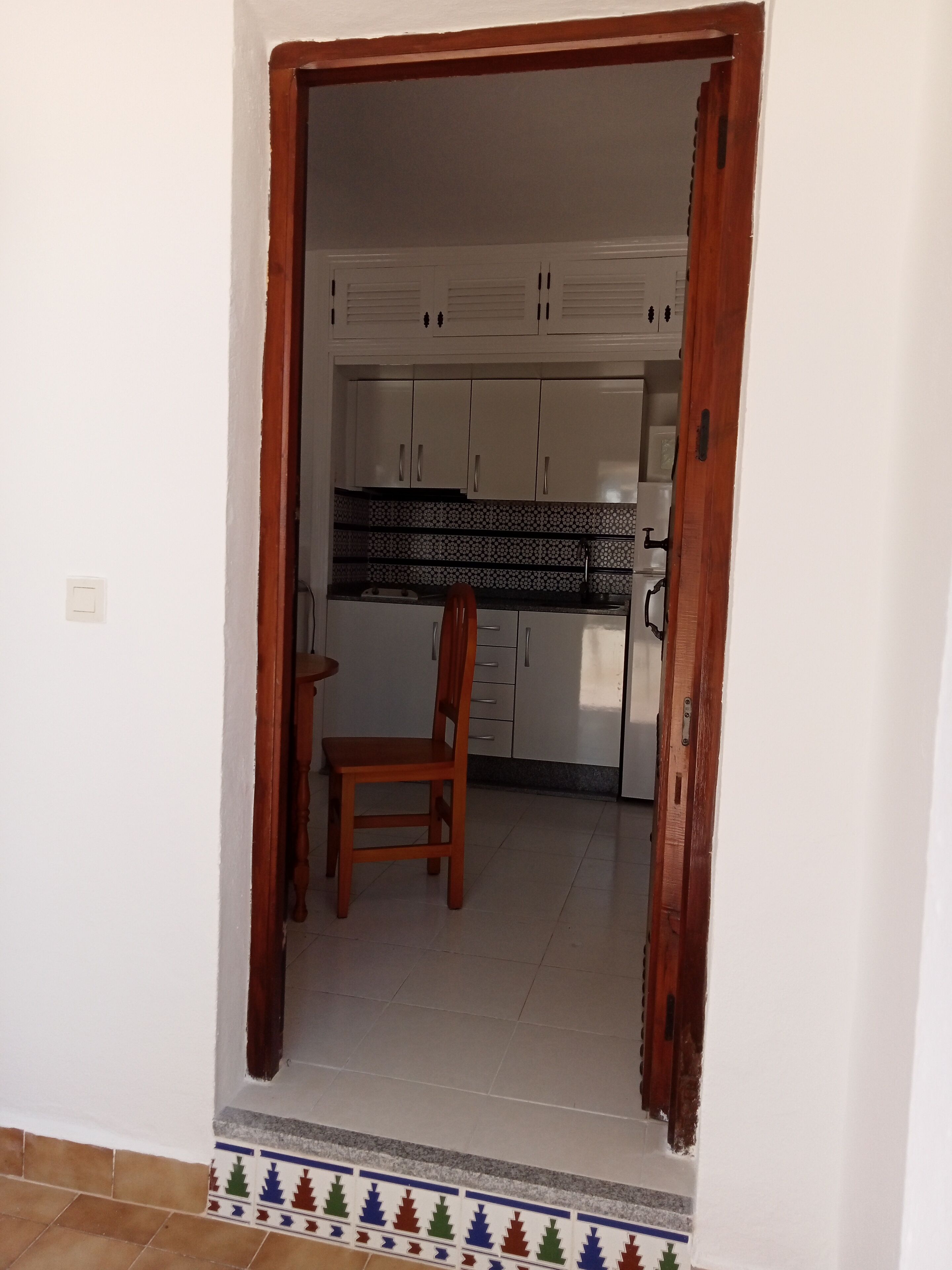Une porte qui mène à une cuisine avec des armoires blanches, une chaise en bois et un mur recouvert de carreaux.