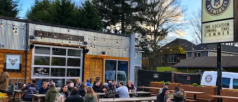 An outdoor seating area with wooden tables and benches, a sign indicating a second location coming this summer, and a building with a large window and a logo.