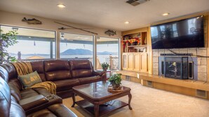 A living room with a leather couch, a coffee table, a fireplace, a TV, and large windows overlooking a body of water.