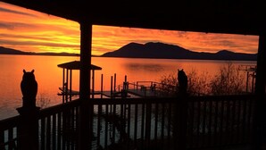 A sunset view from a covered deck with a pier extending into a calm lake, surrounded by mountains.