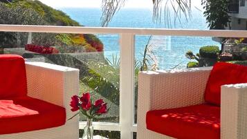Un balcon agrémenté de meubles en osier et de coussins rouges, offrant une vue sur un paysage côtier avec la mer et des bâtiments en toile de fond.