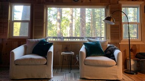 A cozy cabin interior with two white armchairs, a small table, and a large window offering a view of tall trees.