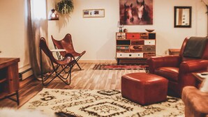 A living room with a wooden floor, a red leather sofa, a wooden coffee table, a red leather armchair, a wooden side table, a small wooden cabinet, and a large rug. - Mountain Retreat Condo (Kremmling)