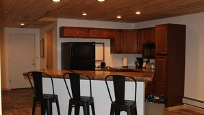 A kitchen with wooden cabinets, a black refrigerator, a microwave, and a coffee maker. There are three black bar stools and a trash can. - Mountain Retreat Condo (Kremmling)