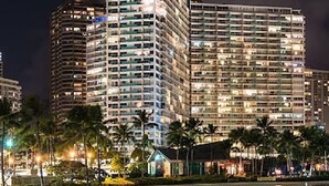 A high-rise hotel with multiple floors, a well-lit facade, and a surrounding area with palm trees and a body of water. - Ocean & Lagoon Views.  Don't miss out on our last-minute discounted rate offer! (Honolulu)