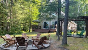 A circular seating area with Adirondack chairs and a fire pit, surrounded by a wooded area with a house in the background. - Serene Setting on the Platte River (Honor)