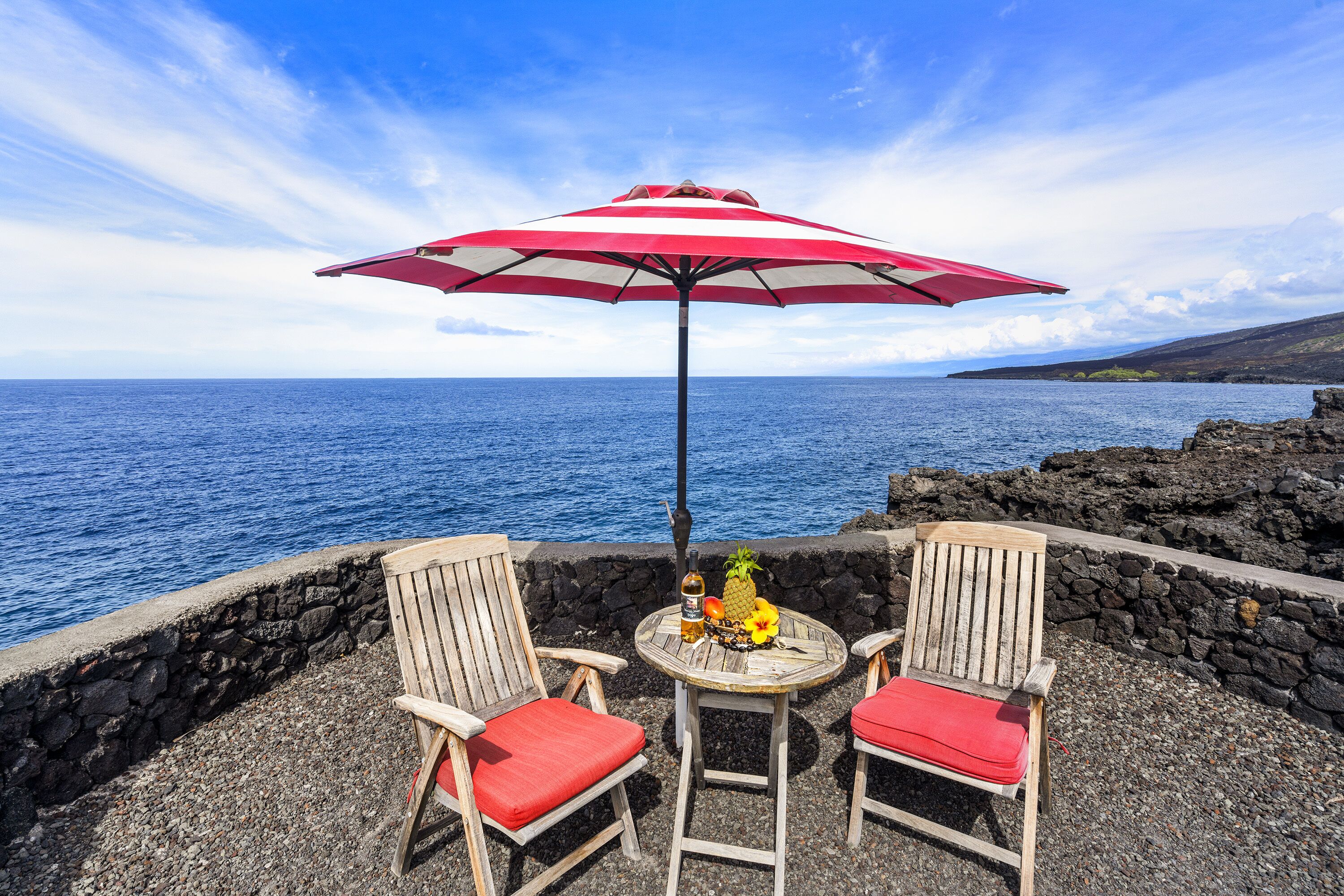 Eine Terrasse am Meer mit zwei Holzstühlen, einem Tisch und einem Sonnenschirm, mit Blick auf den Ozean.