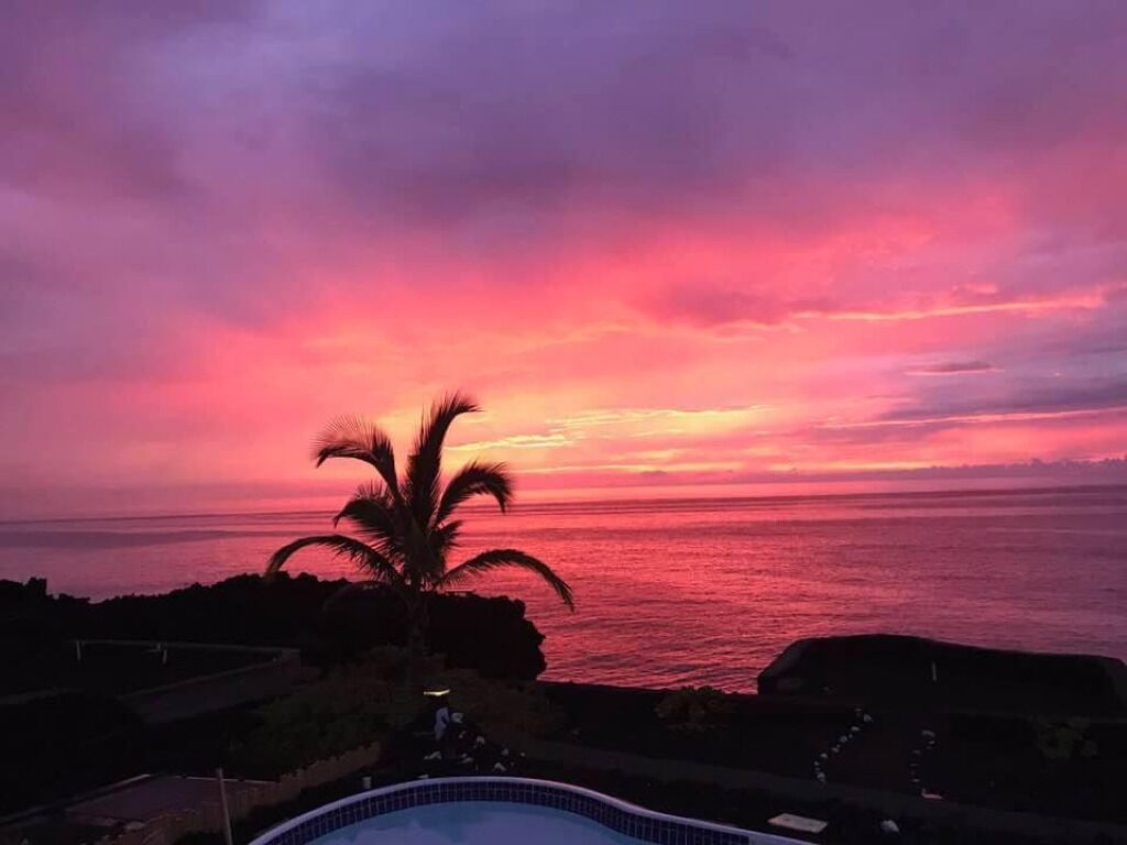 A sunset view over a body of water with a palm tree and a pool in the foreground.