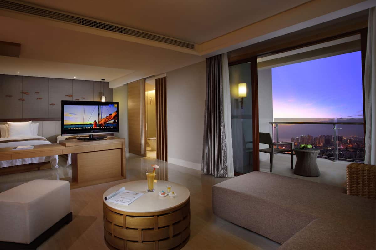 A modern hotel room with a large TV, a balcony with a city view, a dining area, and a cozy seating space.