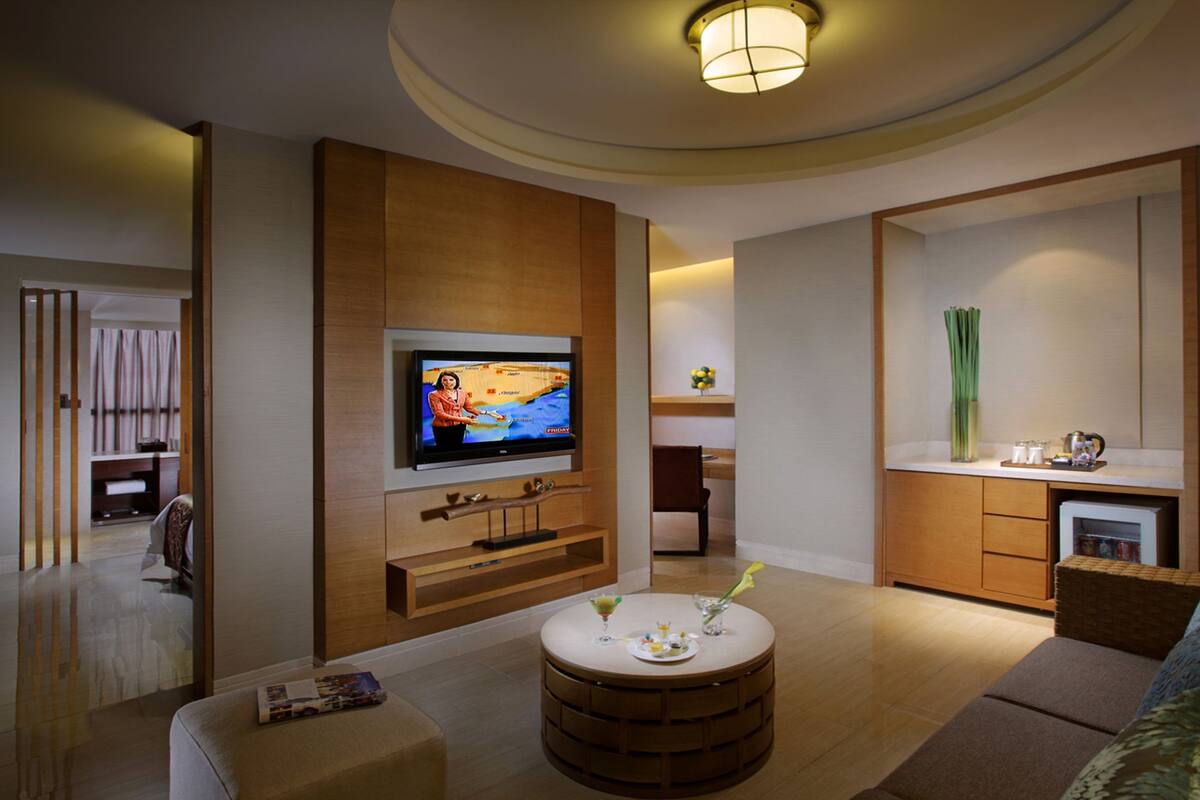 A modern living room with a flat-screen TV, wooden furniture, a round coffee table, and a kitchen area with a sink and cabinets.