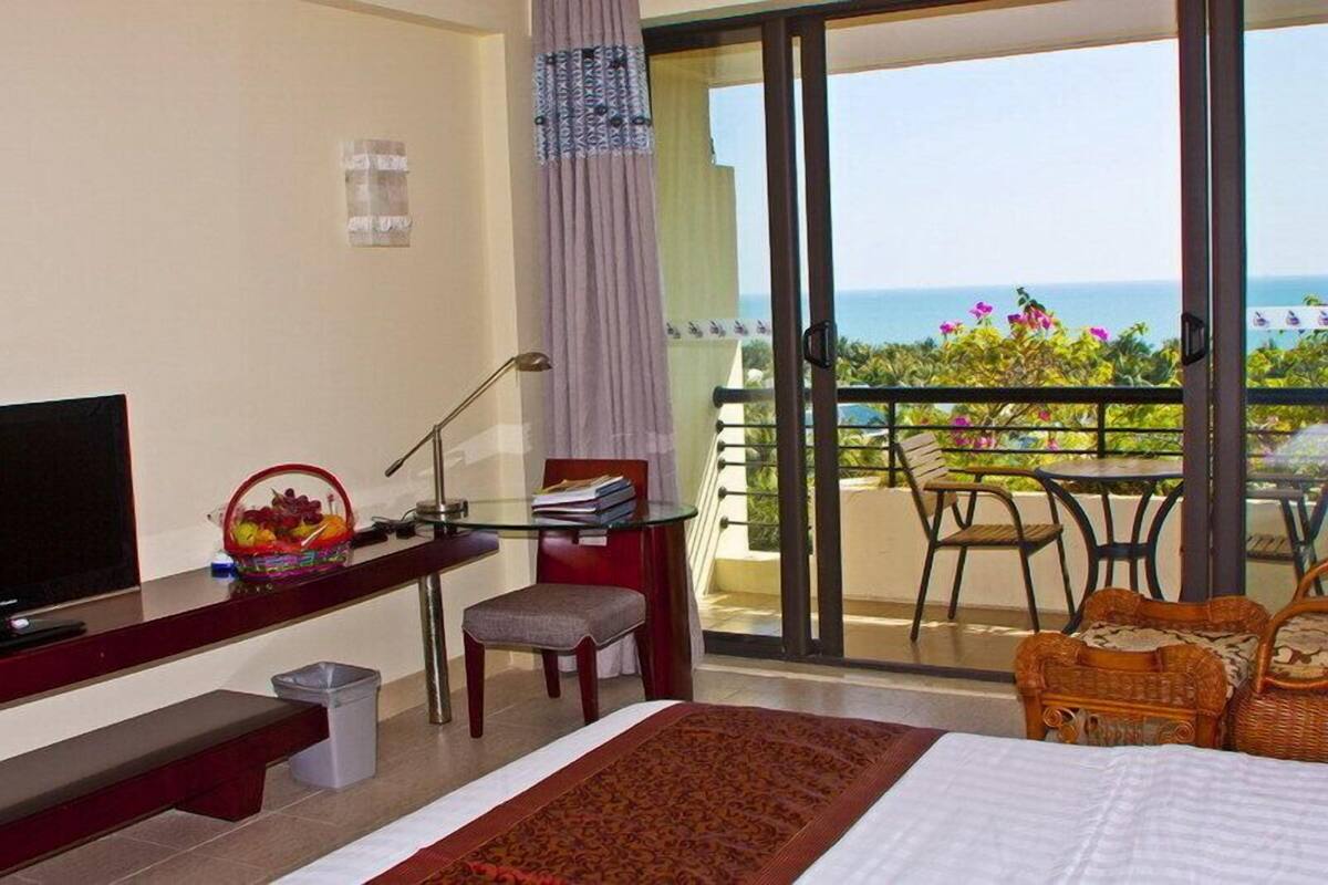 Quarto de hotel com cama, escrivaninha com televisão, cadeira, um cesto de lixo pequeno e varanda com vista para o mar.