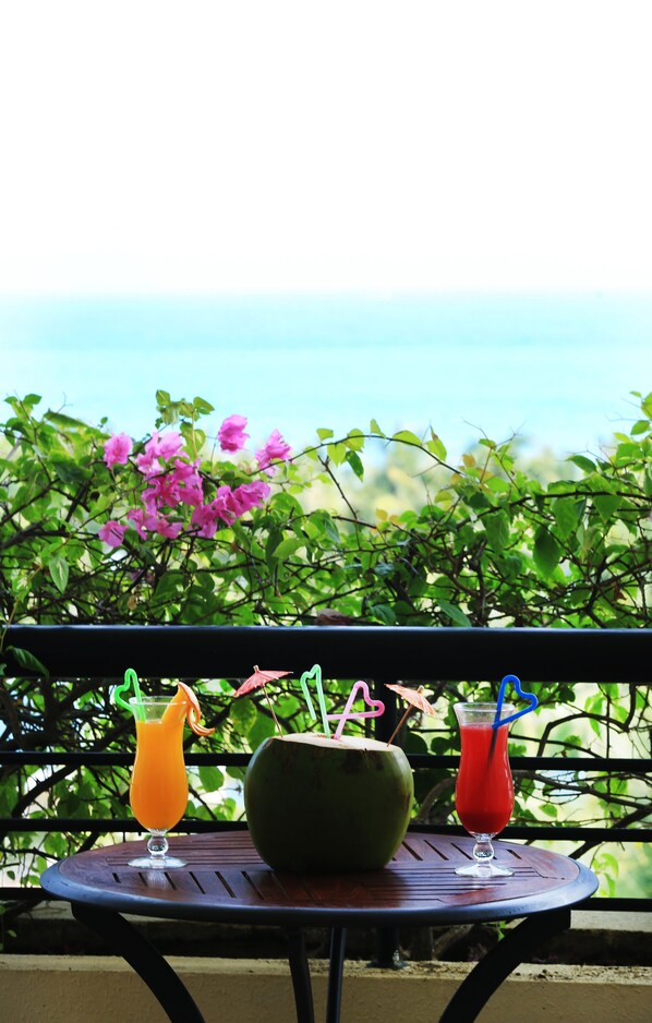 Una mesa con dos bebidas y un coco en un balcón con vista al mar y flores rosadas.