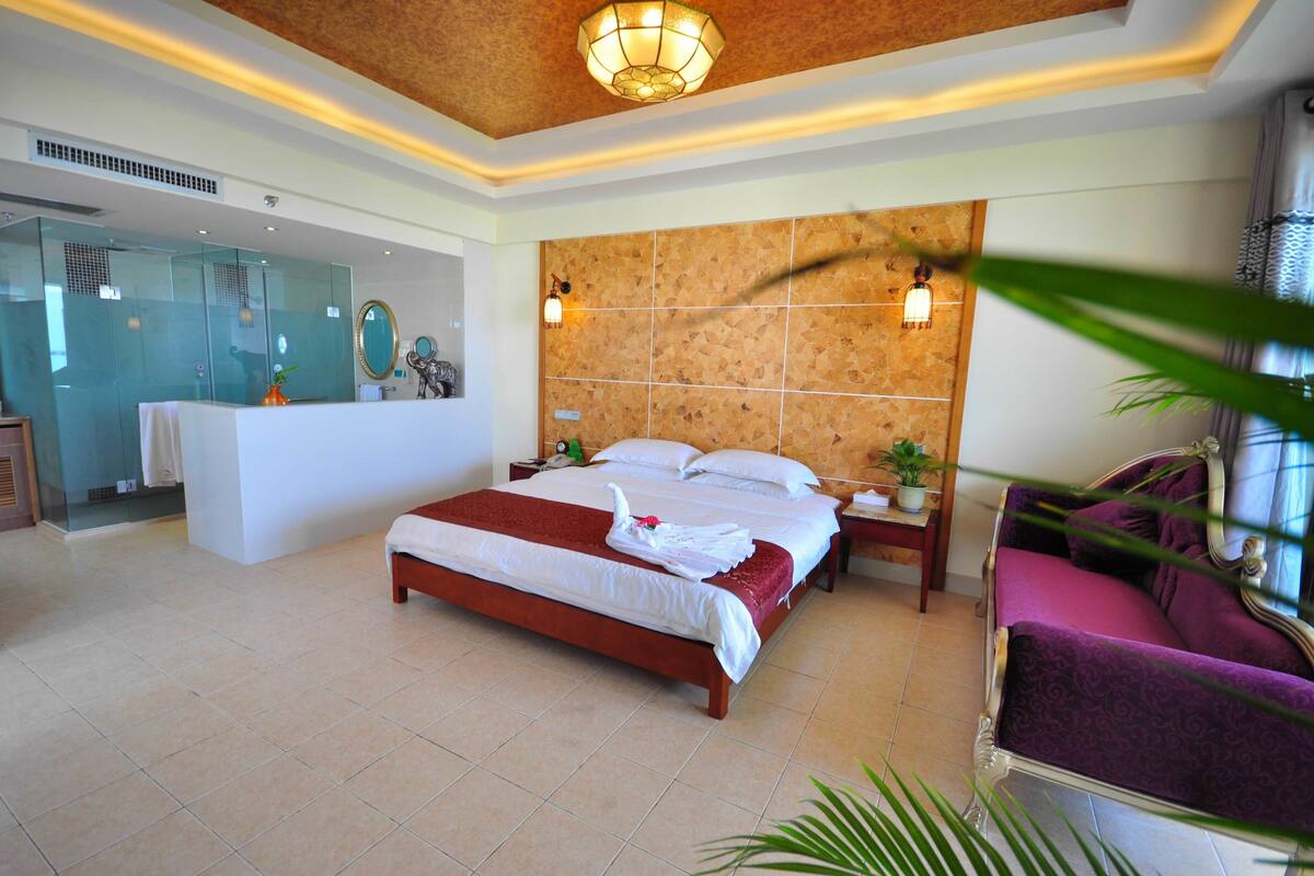 Quarto de hotel com uma cama grande, toucador com espelho, área de estar com sofá roxo e luminária decorativa no teto.