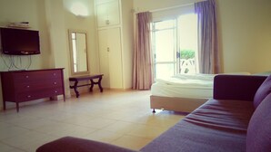 A bedroom with a bed, a sofa, a dresser, a TV, a window with curtains, and a mirror. - Kibbutz Tiratzvi - Country Lodging (Emek HaMaayanot)