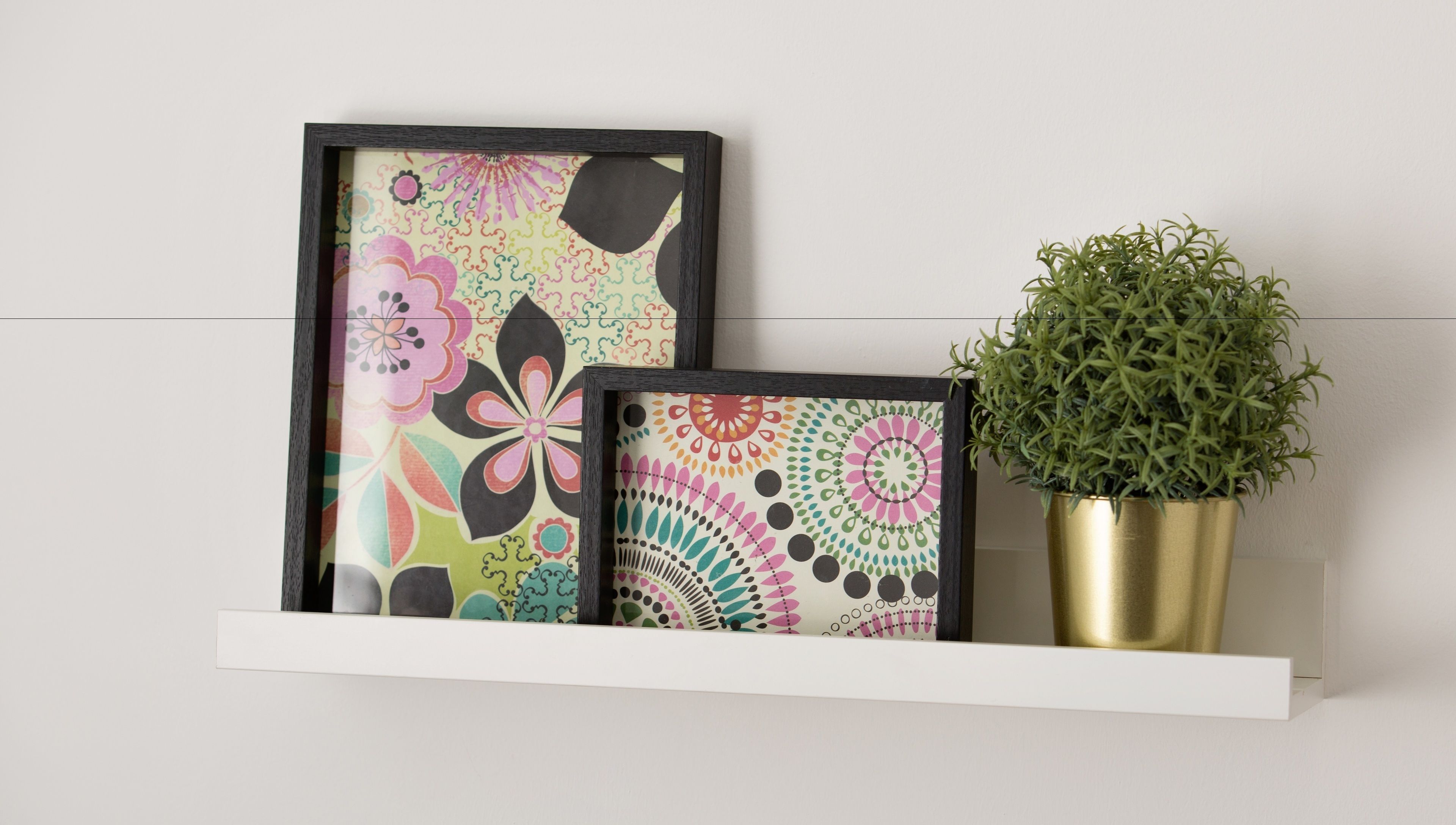 Two framed artworks with a plant in a gold pot on a white shelf.