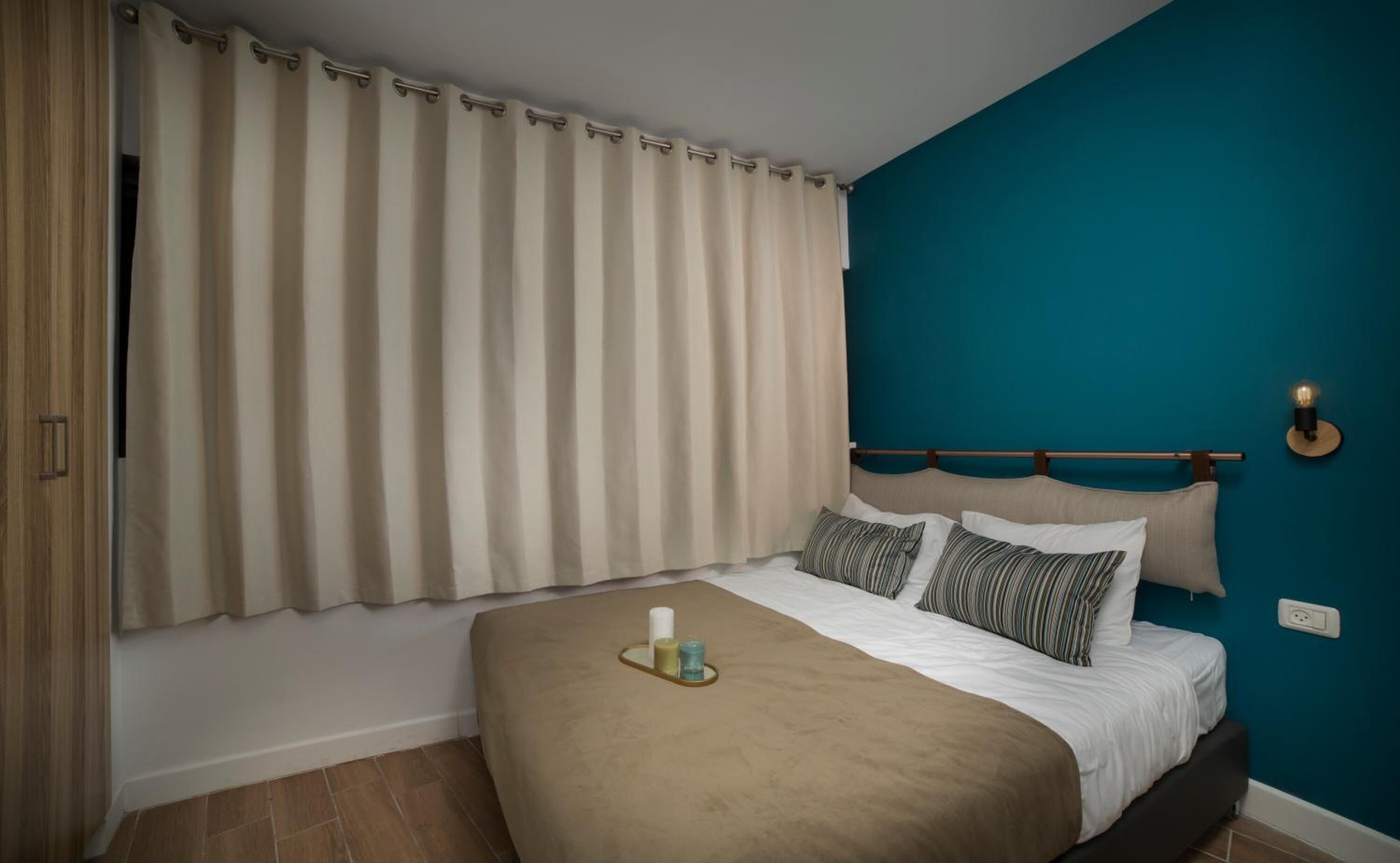 Un dormitorio con cama, cortinas, cabecera, mesita de noche con velas y un enchufe en la pared.