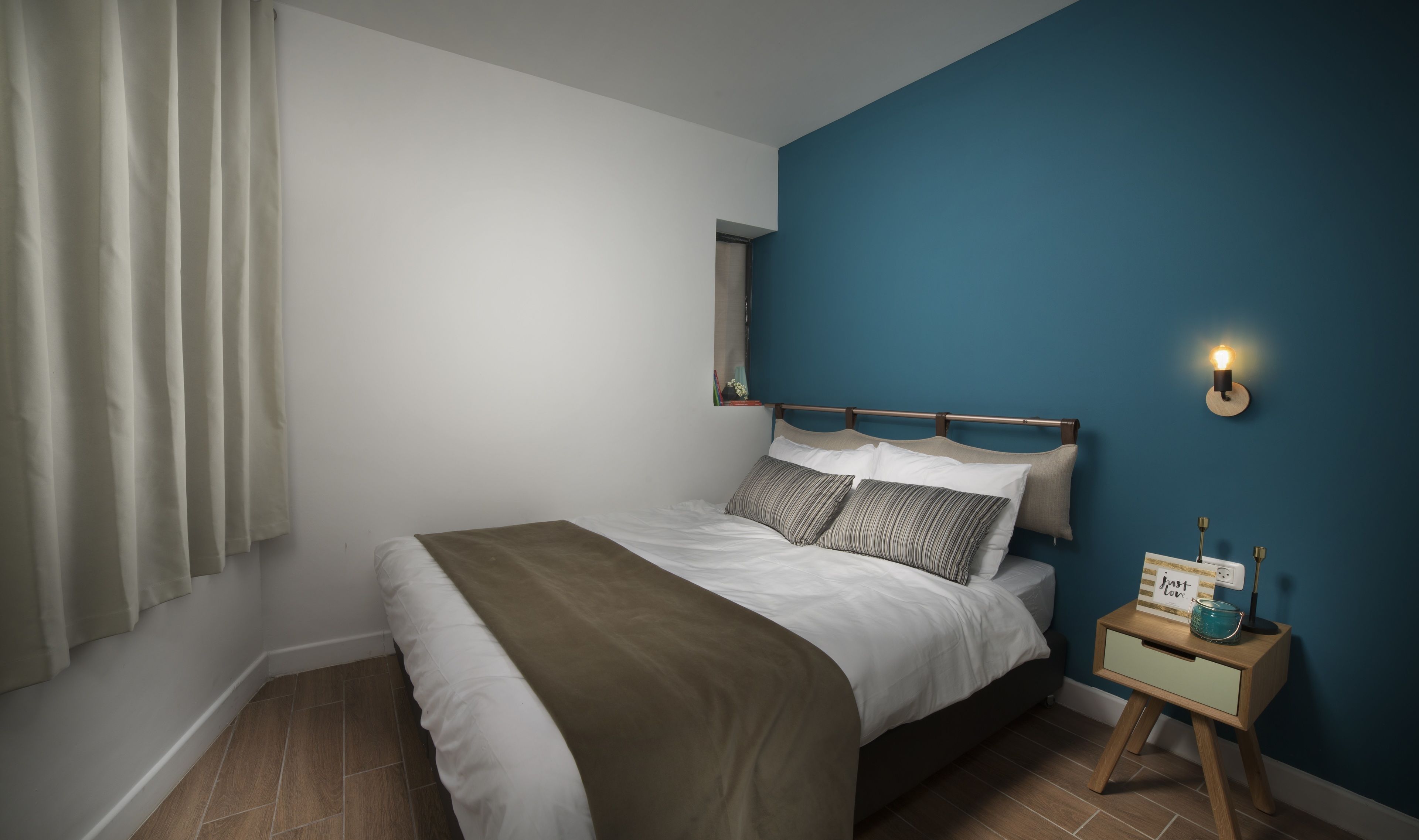 Un dormitorio con cama, mesita de noche, lámpara de pared y ventana con cortinas.