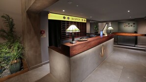 A reception area with a wooden reception desk, a reception sign, a lamp, and a staircase in the background. - Diana Hotel (Haifa)