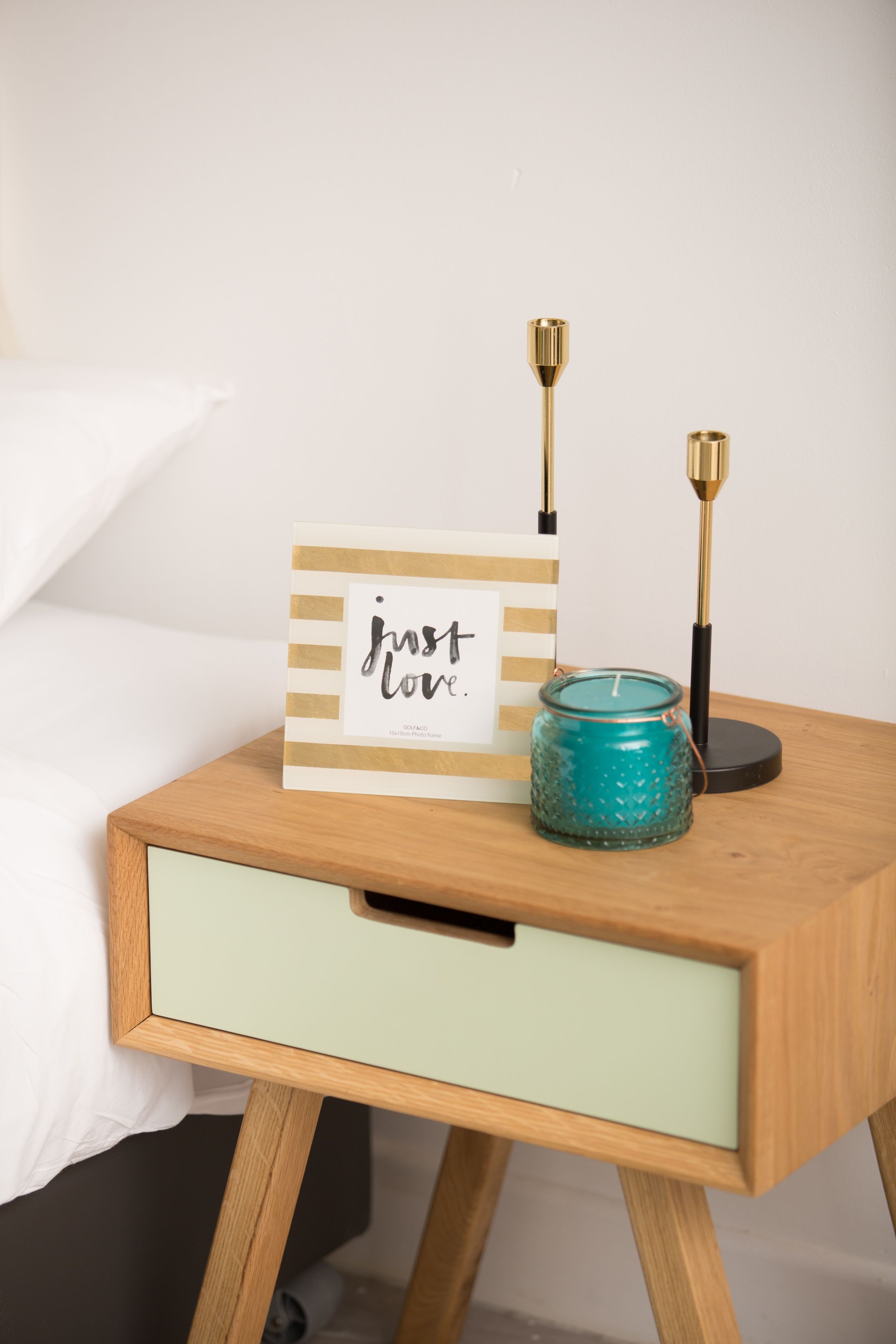A bedside table with a framed sign, a teal candle jar, and two candlesticks.