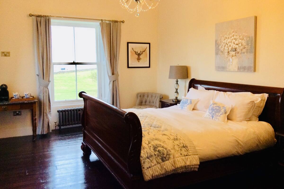A bedroom with a large bed, a chandelier, a window with curtains, a radiator, a desk with a lamp, and a framed picture on the wall.
