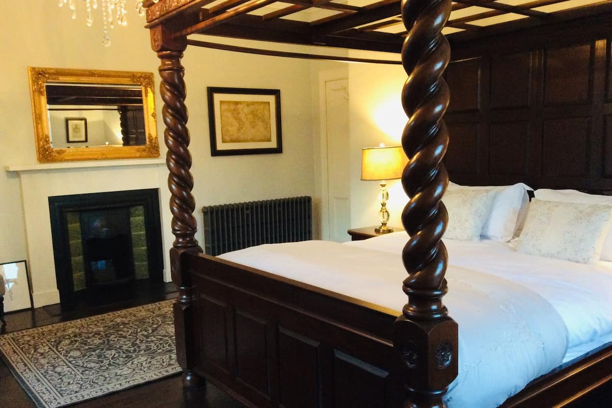 A four-poster bed with a dark wooden frame and white bedding, a fireplace with a mirror above it, and a framed picture on the wall.