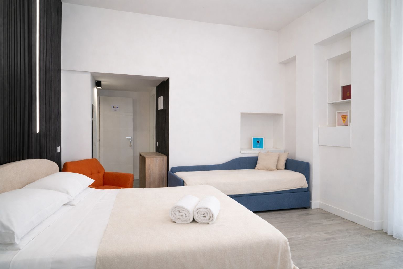 Una habitación de hotel con dos camas, una silla, una mesita y un estante con objetos.