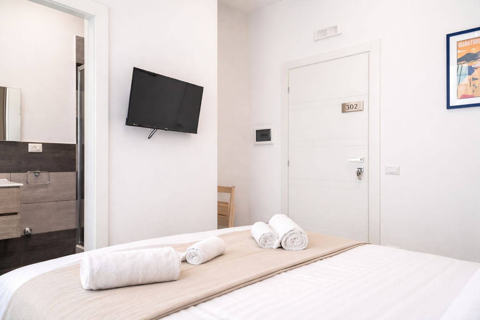A hotel room with a bed, a TV mounted on the wall, a door with a number, and a bathroom visible through an open door.