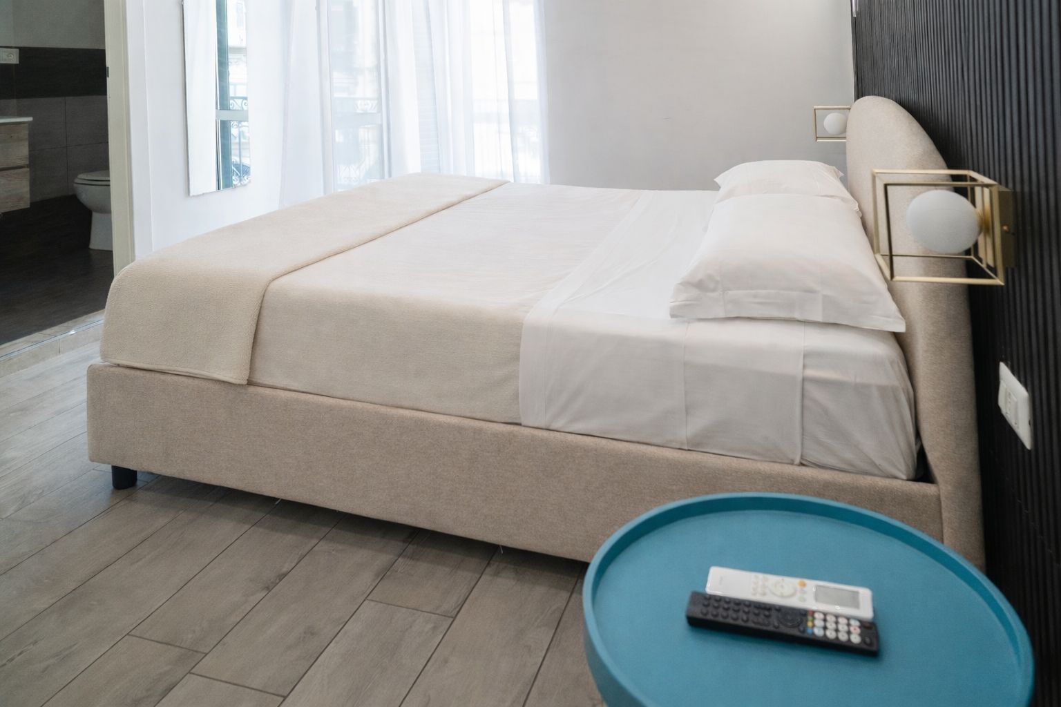 A modern hotel room with a large bed, a bedside table with a remote control, and a view of the city through large windows.