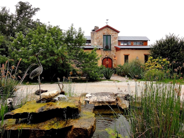 A rustic stone building with a red door and a small tower, surrounded by lush greenery and a pond.