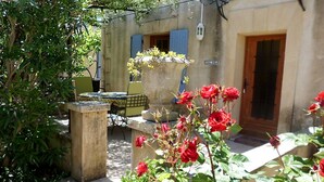 Um pátio com mesa e cadeiras, um vaso grande com flores e um prédio com porta e janela.