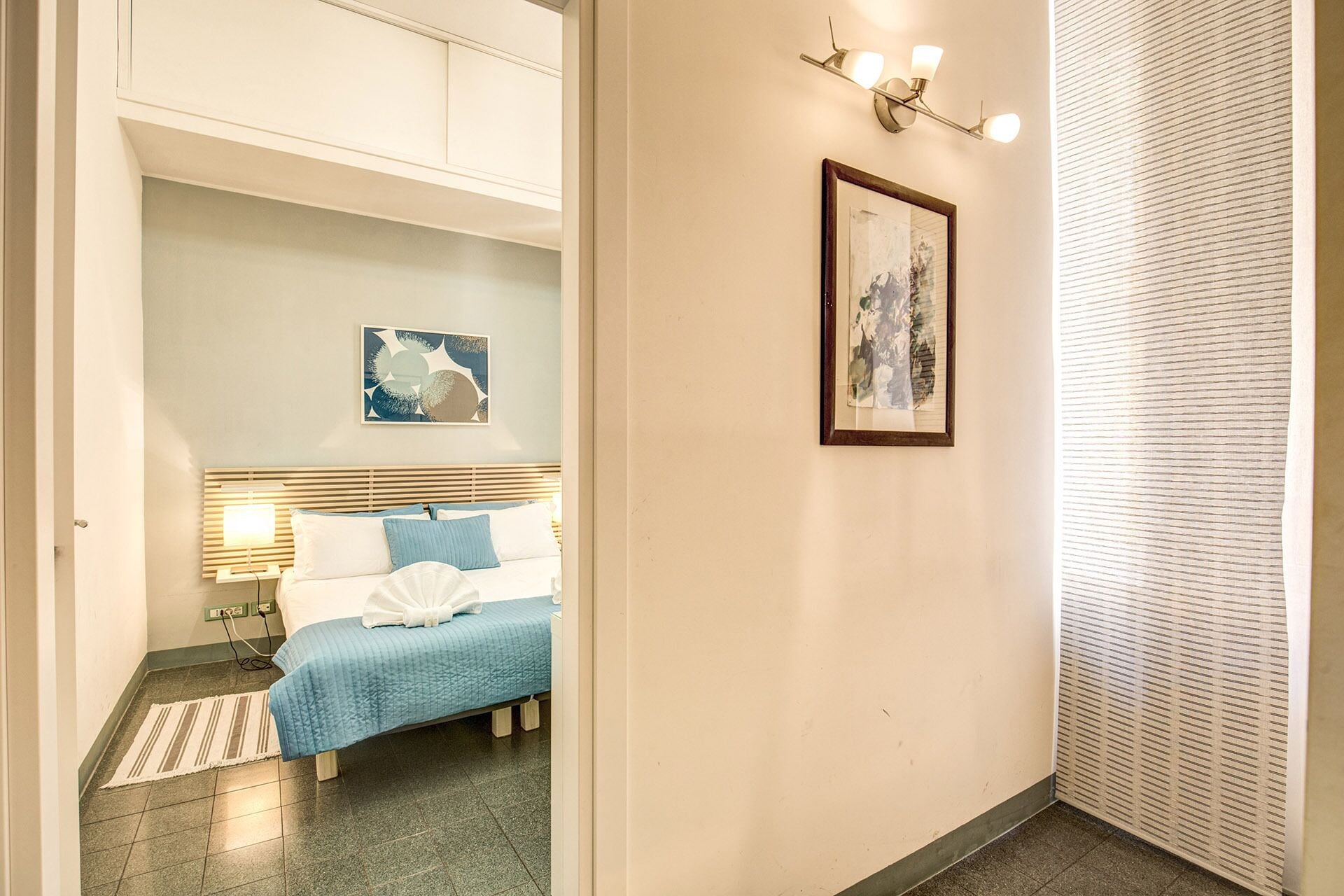 Una habitación de hotel con una cama, una mesita de noche, una lámpara de pared e imagen en un marco.