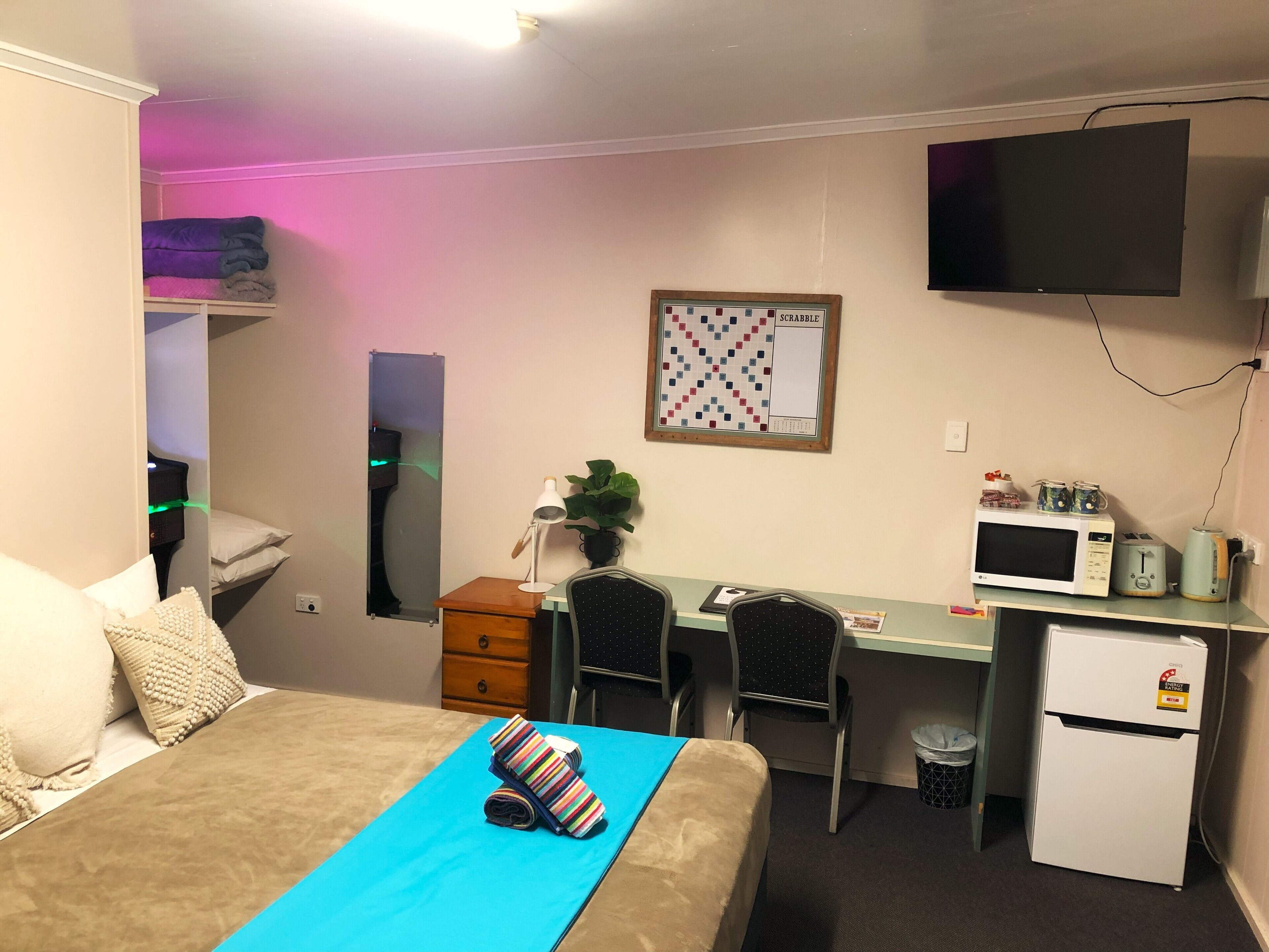 Habitación compacta con cama, escritorio, silla, televisor, microondas y un pequeño cesto de basura.
