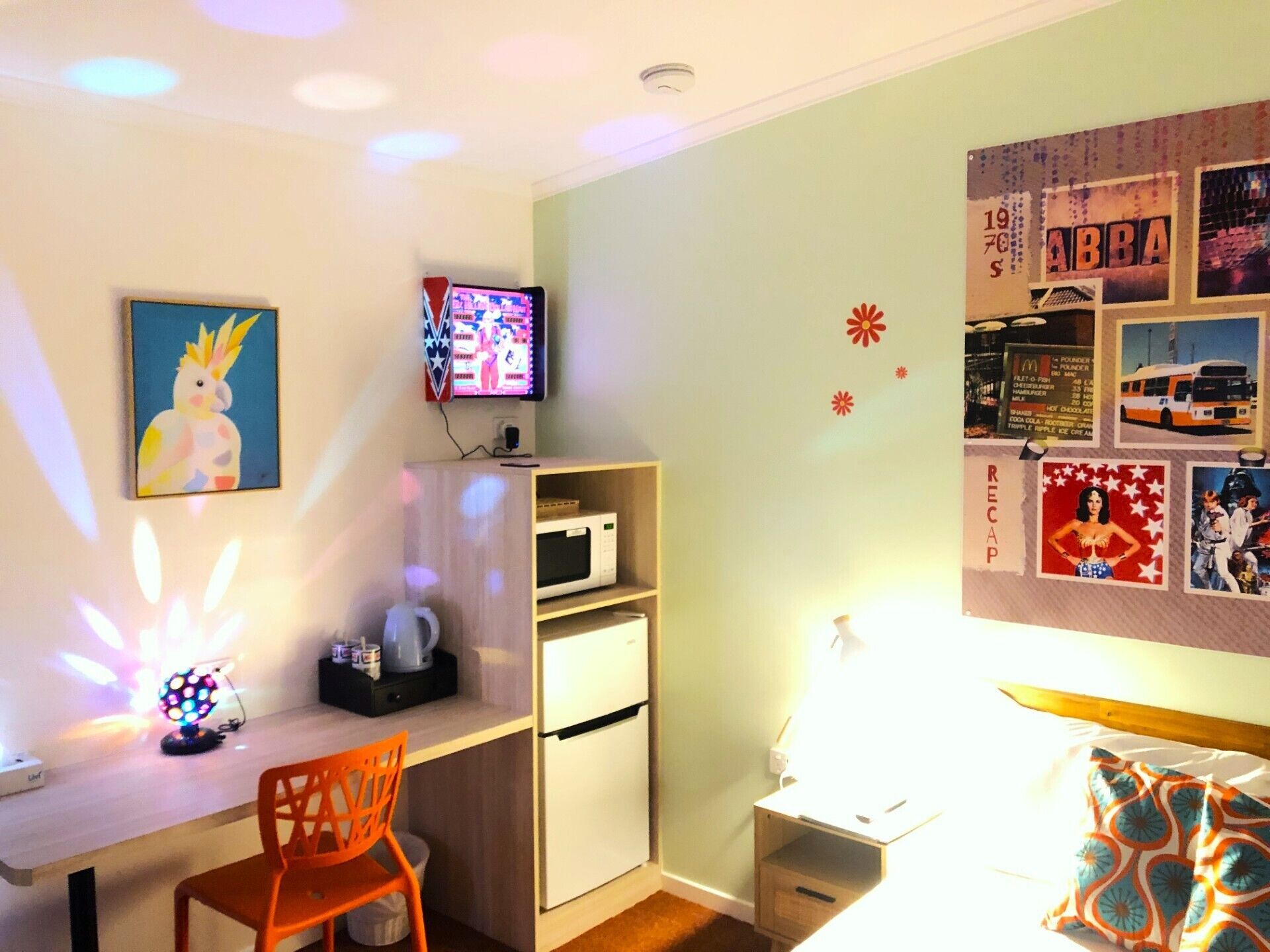 A small, well-lit room with a desk, a chair, a microwave, and a bed. There are wall decorations and a television on the wall.