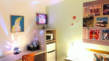 A small, well-lit room with a desk, a chair, a microwave, and a bed. There are wall decorations and a television on the wall.