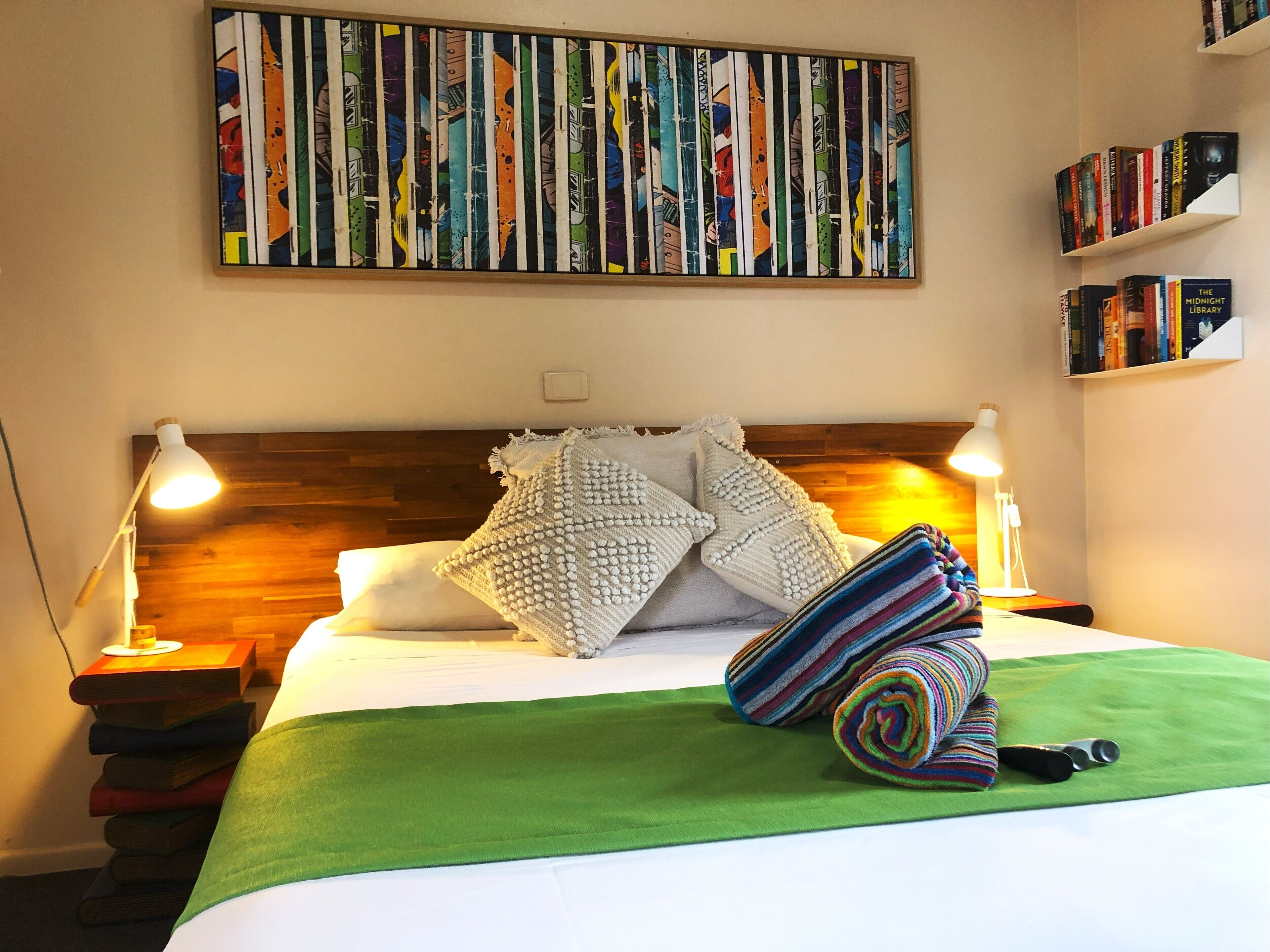 A neatly made bed with a green bedspread, two white pillows, and a colorful striped blanket. There are two bedside lamps, a wooden headboard, and a bookshelf with books.