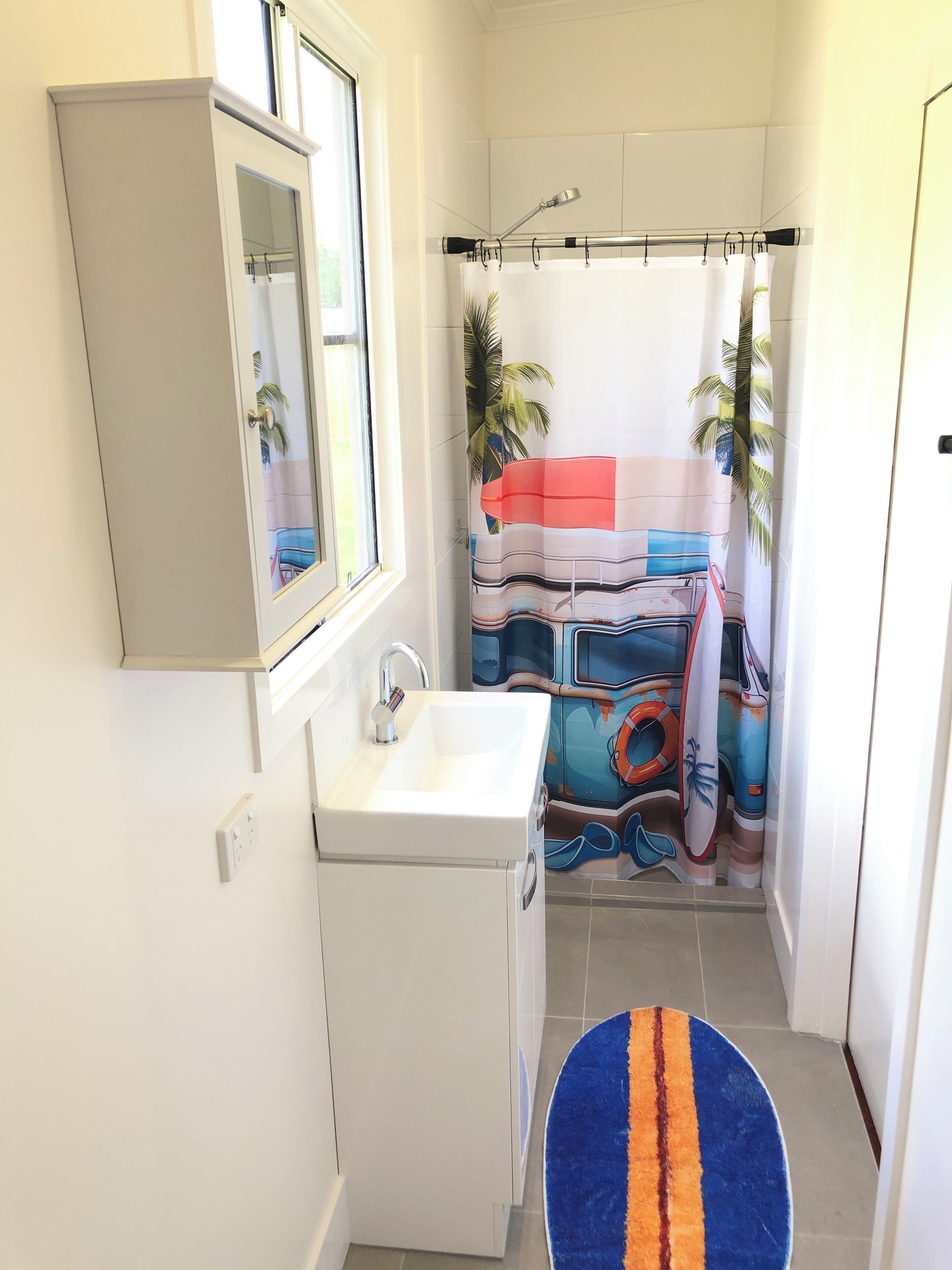 Un baño con un lavamanos blanco, un espejo encima, una cortina de ducha con diseño tropical y una alfombra azul y naranja en el suelo.