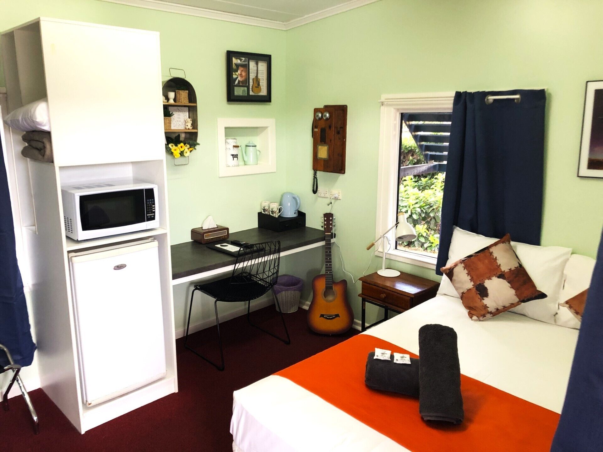 Een kleine kamer met een bed, een bureau met stoel, een gitaar, een klein tafeltje met lamp en een raam met gordijnen.